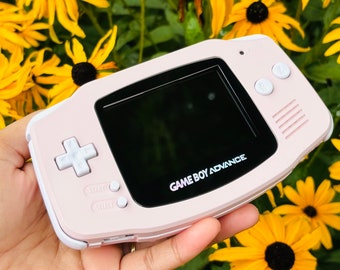May include: A pink Game Boy Advance handheld video game console with a black screen and white buttons. The console is in the hand of a person, with a blurred background of yellow flowers.