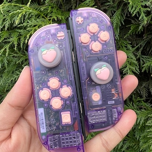 May include: A pair of purple Nintendo Switch Joy-Con controllers with peach-shaped buttons. The controllers have a clear, translucent design, revealing the internal components.