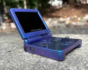 May include: A purple Game Boy Advance SP with a black screen and buttons. The device is sitting on a gray surface.