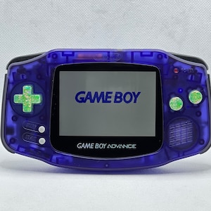 Può includere: Una console per videogiochi portatile Game Boy Advance di colore viola con un pad direzionale verde e bianco. Lo schermo mostra il testo "GAME BOY" e "GAME BOY ADVANCE".