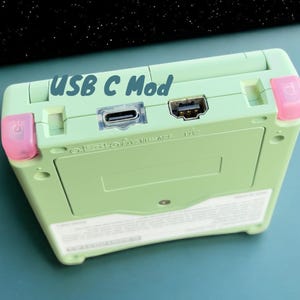 May include: A green Game Boy Advance SP with a USB-C mod. The device has a pink power button and a pink volume button. The text "USB C Mod" is written on the top of the device.