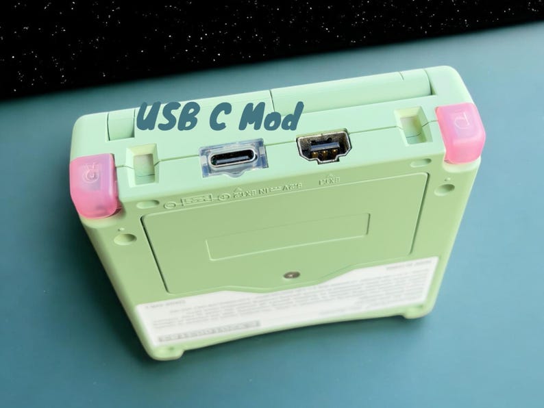 May include: A green Game Boy Advance SP with a USB-C mod. The device has a pink power button and a pink volume button. The text "USB C Mod" is written on the top of the device.