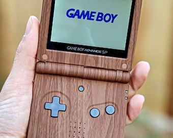 Könnte beinhalten: Ein brauner Game Boy Advance SP mit Holzmaserung. Der Bildschirm zeigt den Text "GAME BOY" in Blau an. Die Knöpfe sind grau.