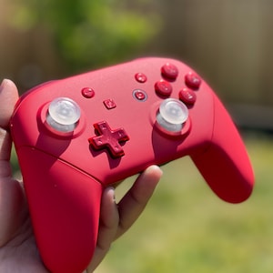May include: A red Nintendo Switch controller with white button covers. The controller has a red plus button and a red home button.