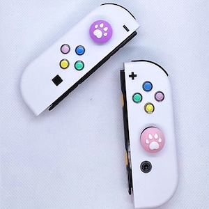May include: Two white Nintendo Switch Joy-Con controllers with colorful buttons and paw print-shaped button caps. The left controller has a purple cap and the right controller has a pink cap.