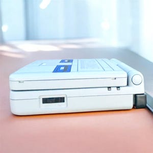 May include: A white handheld gaming console with blue accents. The device is rectangular with a hinged design and a small screen. The console has a slot for game cartridges and a power button. The device is on a pink surface.