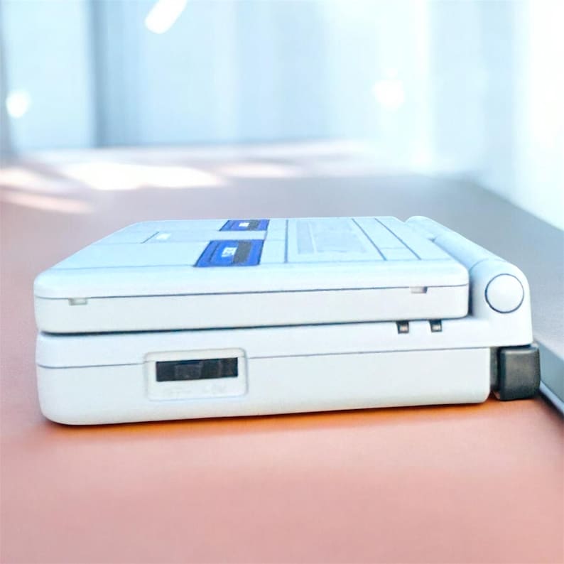 May include: A white handheld gaming console with blue accents. The device is rectangular with a hinged design and a small screen. The console has a slot for game cartridges and a power button. The device is on a pink surface.