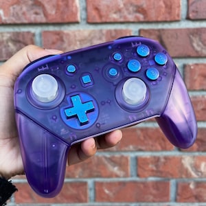 May include: A purple and blue translucent Nintendo Switch controller with white buttons and a blue directional pad.