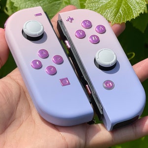 Puede incluir: Un par de mandos Nintendo Switch Joy-Con de color morado y blanco con botones rosas. Los mandos están en un esquema de color degradado, con la mitad superior morada y la mitad inferior blanca.