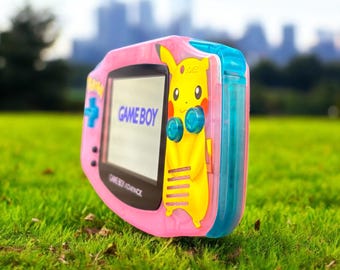 May include: A pink Game Boy Advance handheld console with a Pikachu design. The screen displays "GAME BOY" in blue text. The console has a blue directional pad and buttons, and a yellow Pikachu character is on the side.