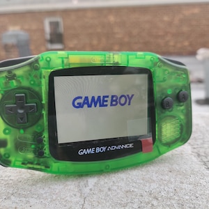 Pode incluir: Um console de jogos portátil Game Boy Advance verde com uma carcaça de plástico transparente. A tela exibe o texto "GAME BOY" em azul.