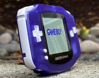 May include: A purple Game Boy Advance handheld video game console with a white faceplate and white buttons. The screen displays the text "GAME BOY" in blue.