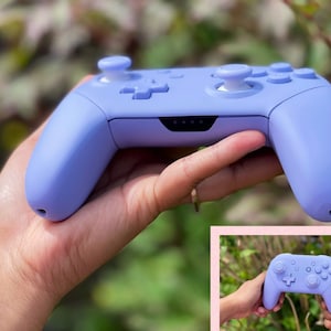 May include: A lavender purple Nintendo Switch controller with white buttons. The controller is held in a hand with a blurred background of green foliage.