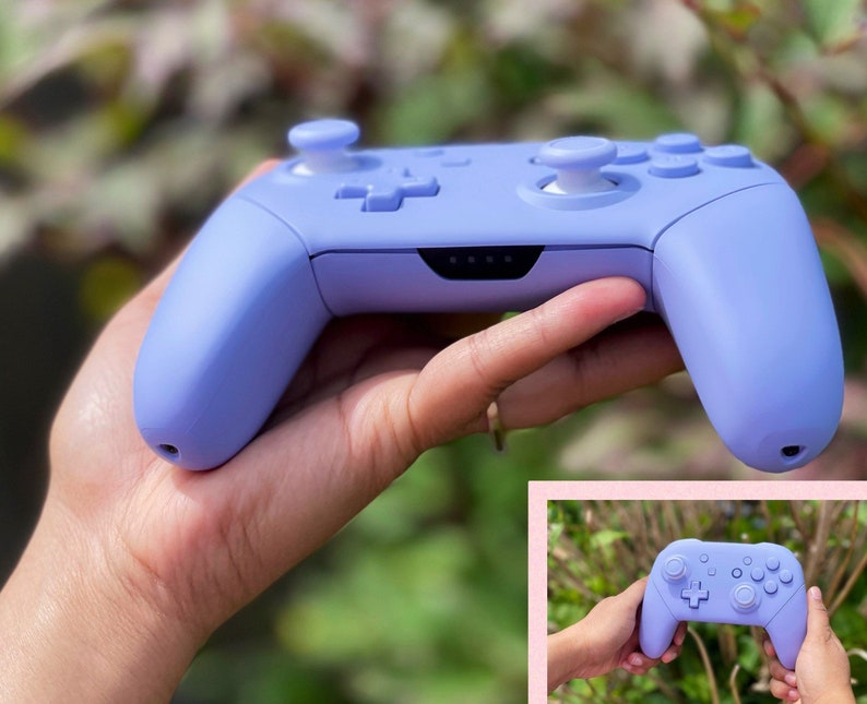 May include: A lavender purple Nintendo Switch controller with white buttons. The controller is held in a hand with a blurred background of green foliage.
