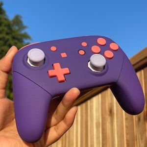 May include: A purple Nintendo Switch controller with pink buttons and white thumbsticks.