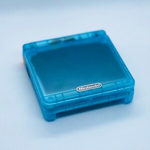 K&ouml;nnte beinhalten: Eine durchsichtige blaue Nintendo Game Boy Advance SP Handheld-Spielkonsole. Die Konsole ist geschlossen und das Nintendo-Logo ist auf der Vorderseite sichtbar.
