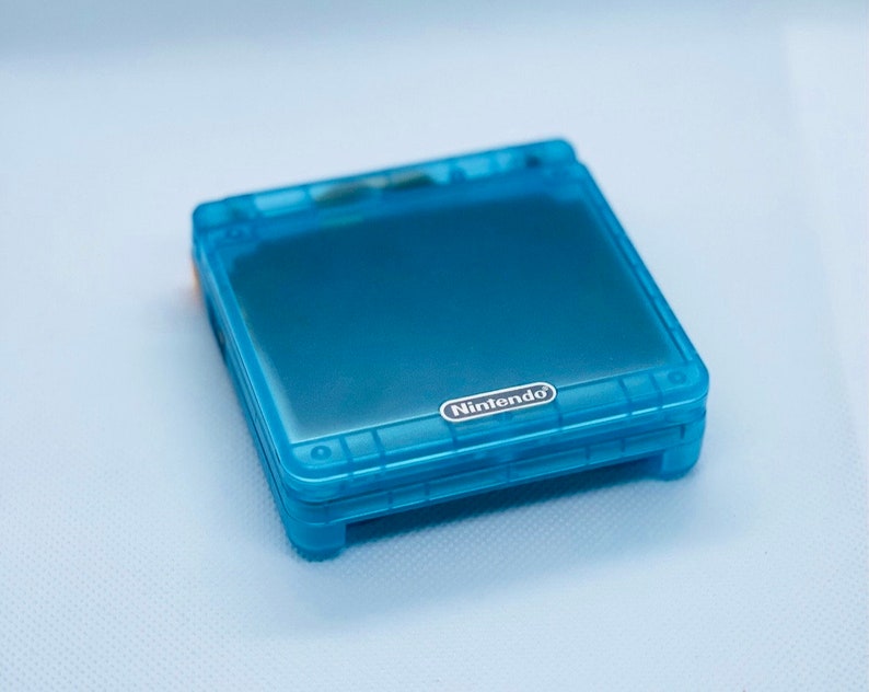 K&ouml;nnte beinhalten: Eine durchsichtige blaue Nintendo Game Boy Advance SP Handheld-Spielkonsole. Die Konsole ist geschlossen und das Nintendo-Logo ist auf der Vorderseite sichtbar.