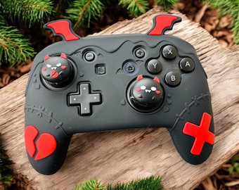May include: A black and red gaming controller with a unique design. The controller features red accents, including bat-wing-shaped details, a broken heart, and a cross-shaped button. The joystick caps have cute devil-themed faces.