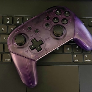 May include: A purple translucent Nintendo Switch controller with black buttons and a black cross button on a laptop keyboard.