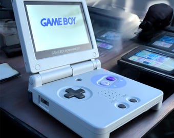 May include: A light gray Game Boy Advance SP handheld game console. The screen displays "GAME BOY" in blue. The console features a directional pad, buttons, and the text "GAME BOY ADVANCE SP".