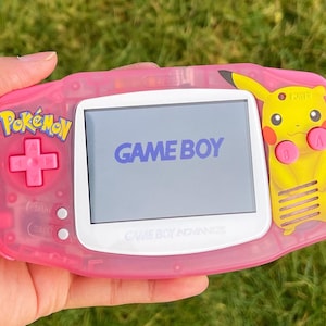Può includere: Una console per videogiochi portatile Game Boy Advance SP rosa con un design Pikachu sul lato destro. La console ha uno schermo bianco con il testo "GAME BOY" visualizzato. La console ha anche un pulsante rosa e un logo "Pokémon" rosa sul lato sinistro.