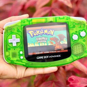 May include: A transparent green Game Boy Advance handheld console displaying the "Pokémon FireRed Version" game. The console has white buttons and a screen displaying a Charizard. The text "GAME BOY ADVANCE" is visible.