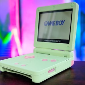 May include: A light green Game Boy Advance SP handheld gaming console with a white screen displaying the text "GAME BOY".