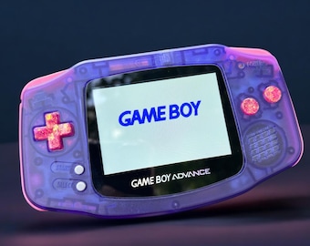 May include: A transparent purple Game Boy Advance handheld console. The screen displays "GAME BOY" in blue. The console has a pink D-pad and buttons, with the words "START" and "SELECT" visible. The text "GAME BOY ADVANCE" is below the screen.