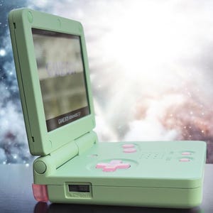 May include: A light green Game Boy Advance SP handheld gaming console with a pink button and a screen displaying the text "GAME BOY ADVANCE SP".