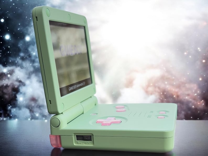 May include: A light green Game Boy Advance SP handheld gaming console with a pink button and a screen displaying the text "GAME BOY ADVANCE SP".