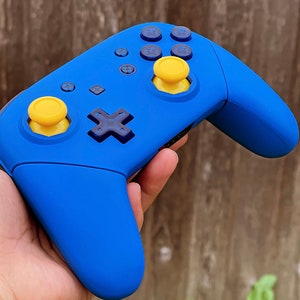 May include: A blue Nintendo Switch controller with yellow and blue buttons. The controller is being held in a hand.