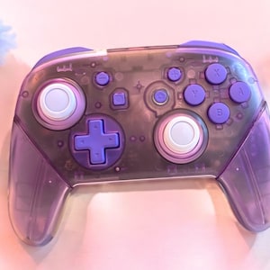 Peut inclure: Une manette de jeu vidéo translucide violette avec des boutons blancs et un bouton en croix violet. La manette a un design en plastique transparent, révélant les composants internes.