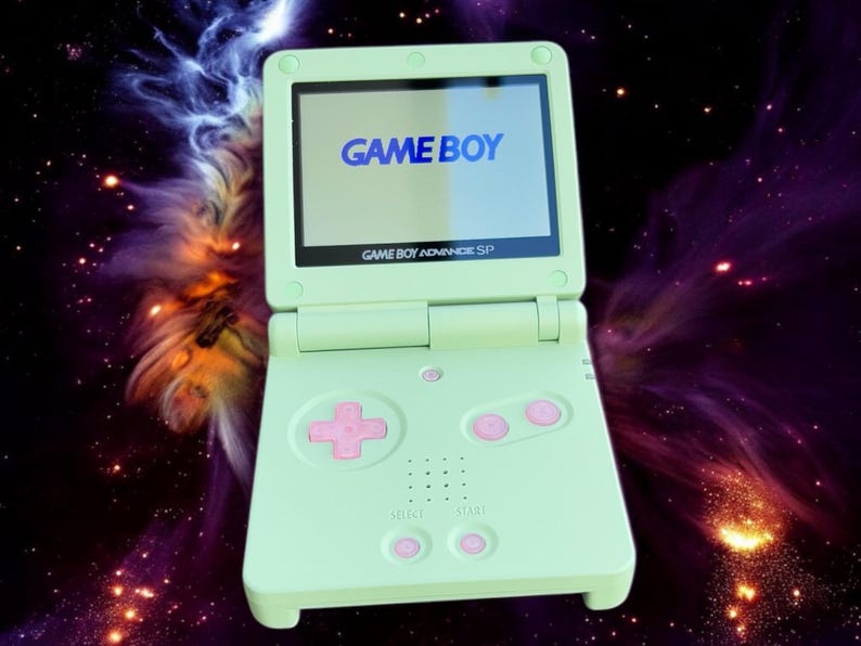 May include: A light green Game Boy Advance SP handheld gaming console with a pink button and a screen displaying the text "GAME BOY".