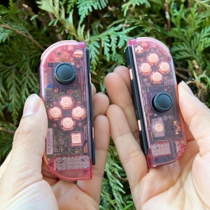 May include: Two clear pink Nintendo Switch Joy-Con controllers with rose gold buttons. The controllers are held in the hands of a person.