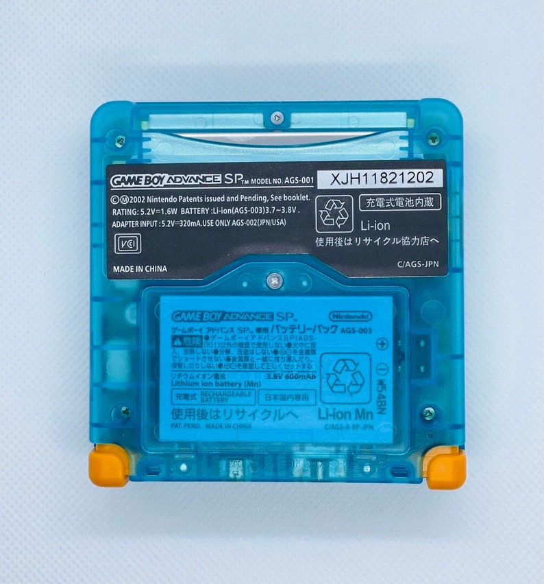 K&ouml;nnte beinhalten: Eine blaue Game Boy Advance SP Handheld-Spielkonsole mit einem schwarz-wei&szlig;en Etikett mit der Aufschrift "GAME BOY ADVANCE SP. MODEL NO. AGS-001 XJH11821202". Das Etikett enth&auml;lt auch Informationen &uuml;ber den Akku, den Adaptereingang und Recyclinghinweise. Die Konsole hat einen gelben Rand um die Kanten.