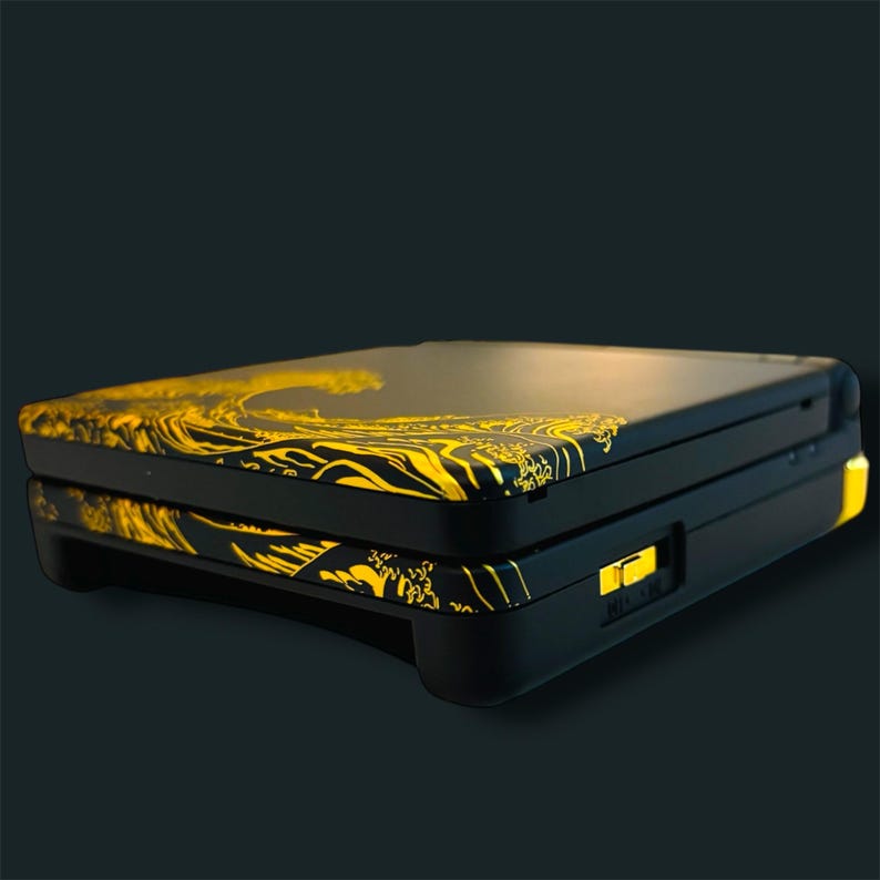 Pode incluir: Consola de jogos port&aacute;til preta com um design de ondas douradas na parte superior e inferior. A consola tem uma forma retangular com cantos arredondados e um bot&atilde;o de ligar/desligar amarelo. O padr&atilde;o de ondas &eacute; uma representa&ccedil;&atilde;o estilizada de ondas oce&acirc;nicas.
