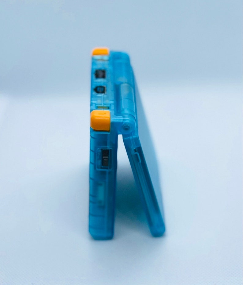 K&ouml;nnte beinhalten: Ein durchsichtiger blauer Game Boy Advance SP mit gelben Kn&ouml;pfen. Die Handheld-Spielkonsole ist geschlossen und steht aufrecht auf einer wei&szlig;en Oberfl&auml;che.