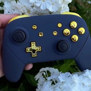 Op de afbeelding: Een marineblauwe Nintendo Switch-controller met gouden knoppen en face buttons. De controller heeft een gouden D-pad en een gouden rand rond de randen.