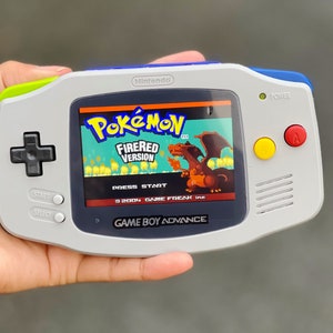 Può includere: Una console per videogiochi portatile Game Boy Advance grigia con un bordo blu e verde. Lo schermo mostra la schermata del titolo del gioco Pokemon FireRed Version con un Charizard rosso.