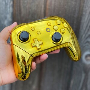 以下が含まれることがあります： 黒いボタンとジョイスティックが付いた金色のニンテンドースイッチコントローラー。コントローラーは光沢のある仕上げで、人の手に持たれています。