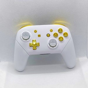 Può includere: Un controller per videogiochi bianco con pulsanti e dettagli dorati. Il controller presenta un D-pad, pulsanti e finiture dorate, con due levette analogiche e un D-pad. Il design generale è elegante e moderno.