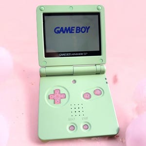 May include: A green Game Boy Advance SP handheld gaming console with a blue "GAME BOY" logo on the screen. The console has pink buttons and a white "SELECT" and "START" button.