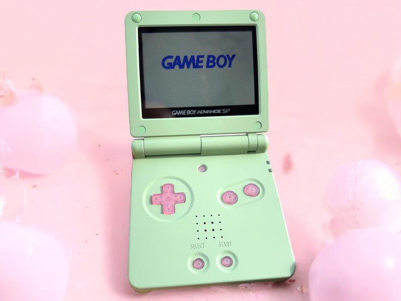 May include: A green Game Boy Advance SP handheld gaming console with a blue "GAME BOY" logo on the screen. The console has pink buttons and a white "SELECT" and "START" button.