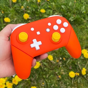May include: A bright orange Nintendo Switch controller with yellow buttons. The controller is held in a hand against a background of green grass and yellow flowers.