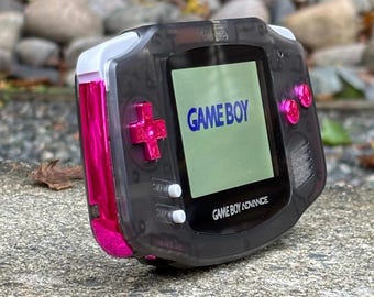 May include: A translucent black Game Boy Advance with a pink trim and white buttons. The screen displays the text "GAME BOY" in blue.