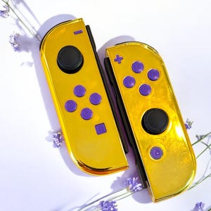 May include: Two gold-colored game controllers with purple buttons and black joysticks. The controllers are set against a white background with purple flowers. The controllers are for a gaming console.