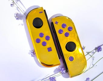 May include: Two gold-colored game controllers with purple buttons and black joysticks. The controllers are set against a white background with purple flowers. The controllers are for a gaming console.