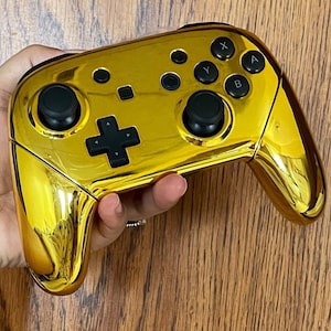 May include: A gold-coloured video game controller with black buttons and joysticks. The controller has a D-pad, X, Y, A, and B buttons. The controller is held in a person's hand.