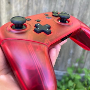 May include: A red translucent video game controller with black buttons and joysticks. The controller has a textured surface and is being held in a hand.