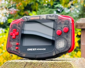 May include: A clear, black Game Boy Advance handheld gaming console with red accents. The console has a transparent shell, revealing the internal components. The buttons are red and the screen is black.